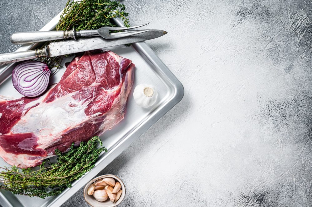 Raw lamb or goat shoulder meat in a baking dish with knife and meat fork.