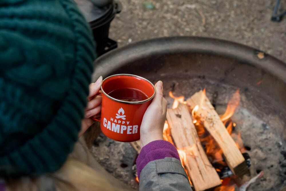 Camping lifestyle concept. Girl holding red enamel mug with tea near campfire. Happy camper